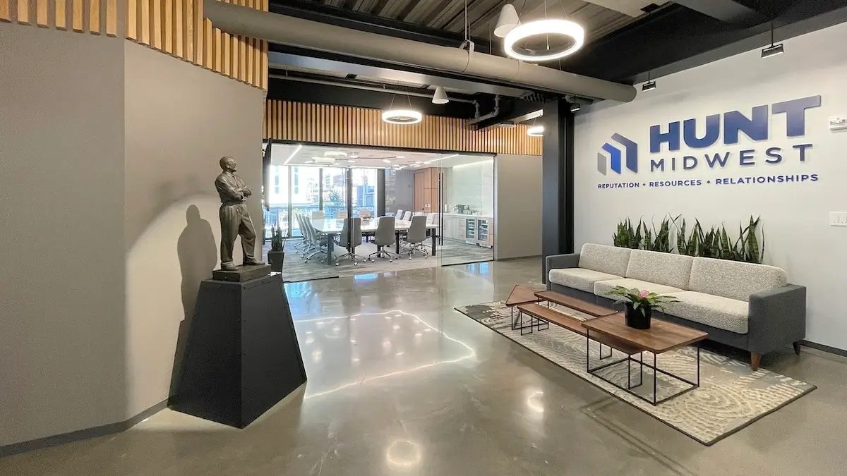 Sleek lobby with gray couch, wooden coffee tables, indoor plants, and a wall logo reading 'Hunt Midwest.' A conference room with glass walls is visible behind.