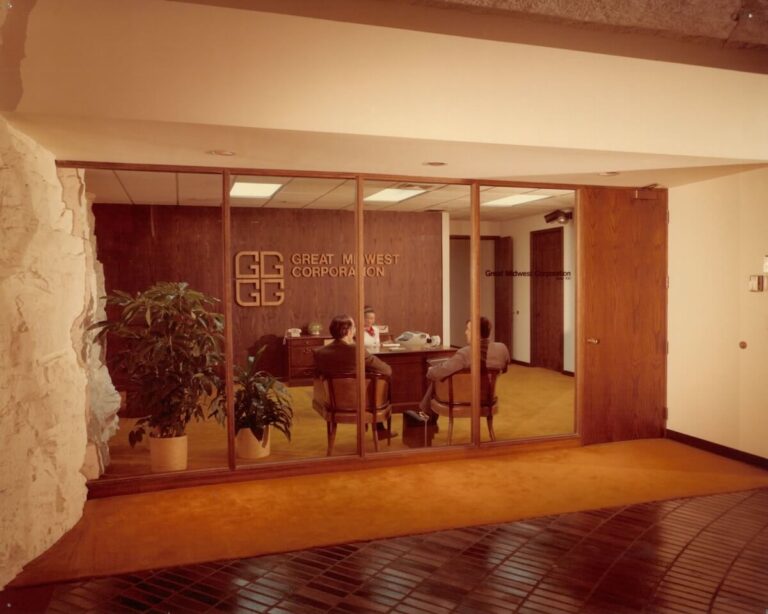 A vintage office space with wood-paneled walls and gold carpeting, featuring the Great Midwest Corporation logo behind a desk where two men are seated in discussion. The office is enclosed by glass walls, with natural rock visible just outside.
