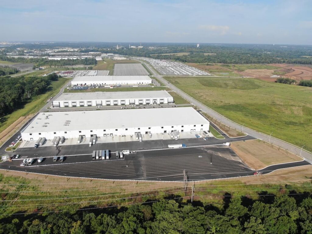 Large white-roofed warehouse buildings with truck docks, parking lots, and surrounding open land extending into the distance.