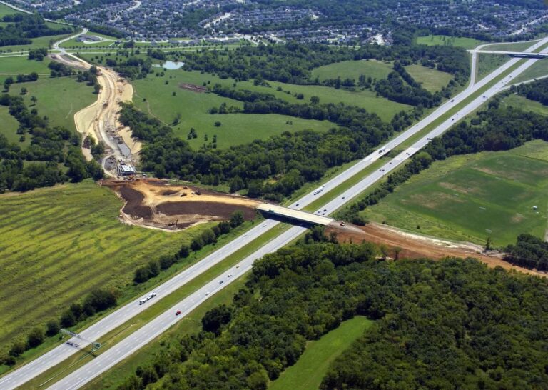 Highway overpass with cars and trucks runs through green fields and trees, while a new road construction project is visible alongside.