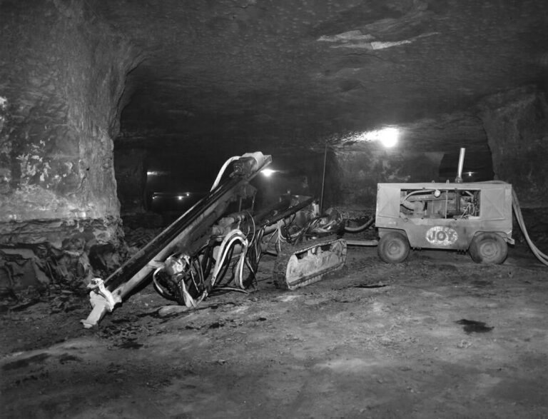 Black and white photo of heavy drilling machinery and a tracked vehicle inside a cavernous underground limestone mine, lit by overhead lamps.