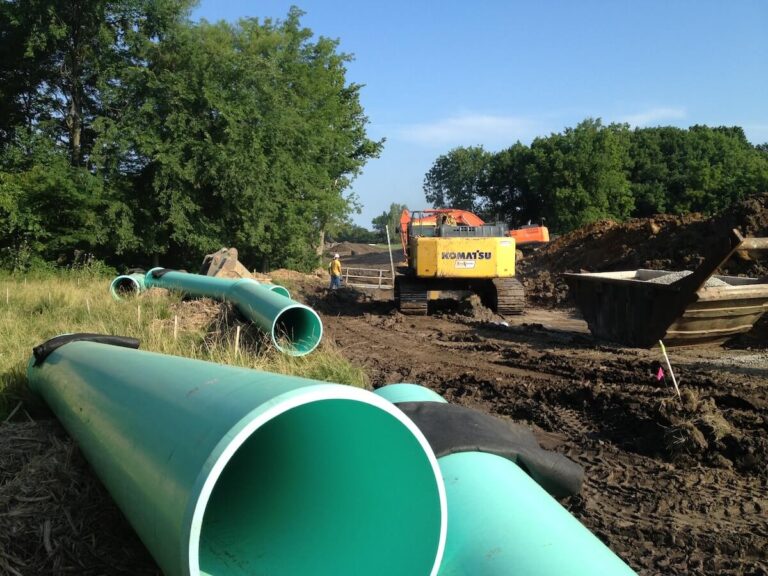 Large green sewer pipes lie on the ground near a Komatsu excavator working in a muddy area with piles of dirt and trees in the background.