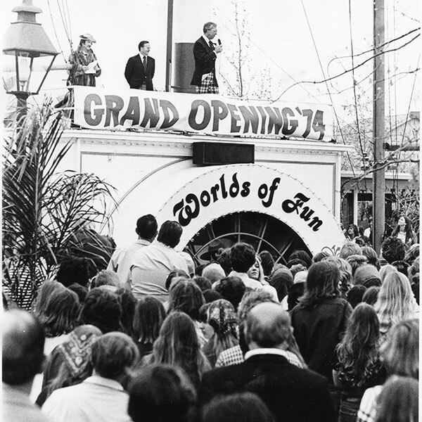 Black-and-white photo of the April 1974 grand opening of Worlds of Fun amusement park. A large crowd gathers below a stage with a banner reading “GRAND OPENING ’74,” as speakers address attendees.