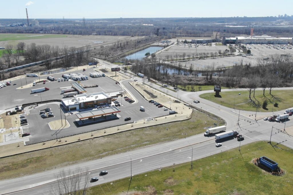 Large fueling station with multiple semi-trucks parked, surrounded by roads, parking lots, and nearby industrial and retail areas.