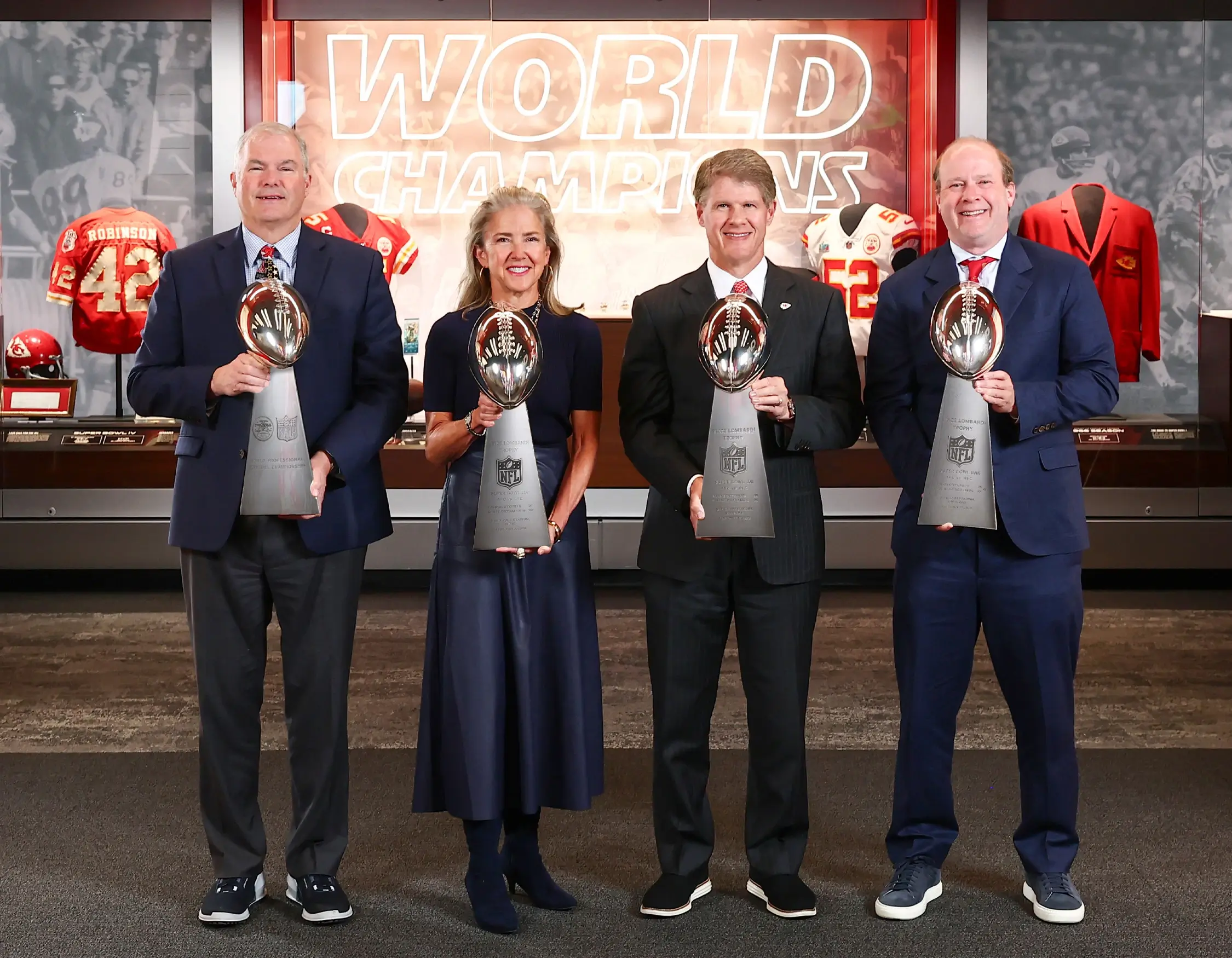 Four people stand side by side holding Vince Lombardi Trophies, with Chiefs jerseys and a 'World Champions' display in the background.