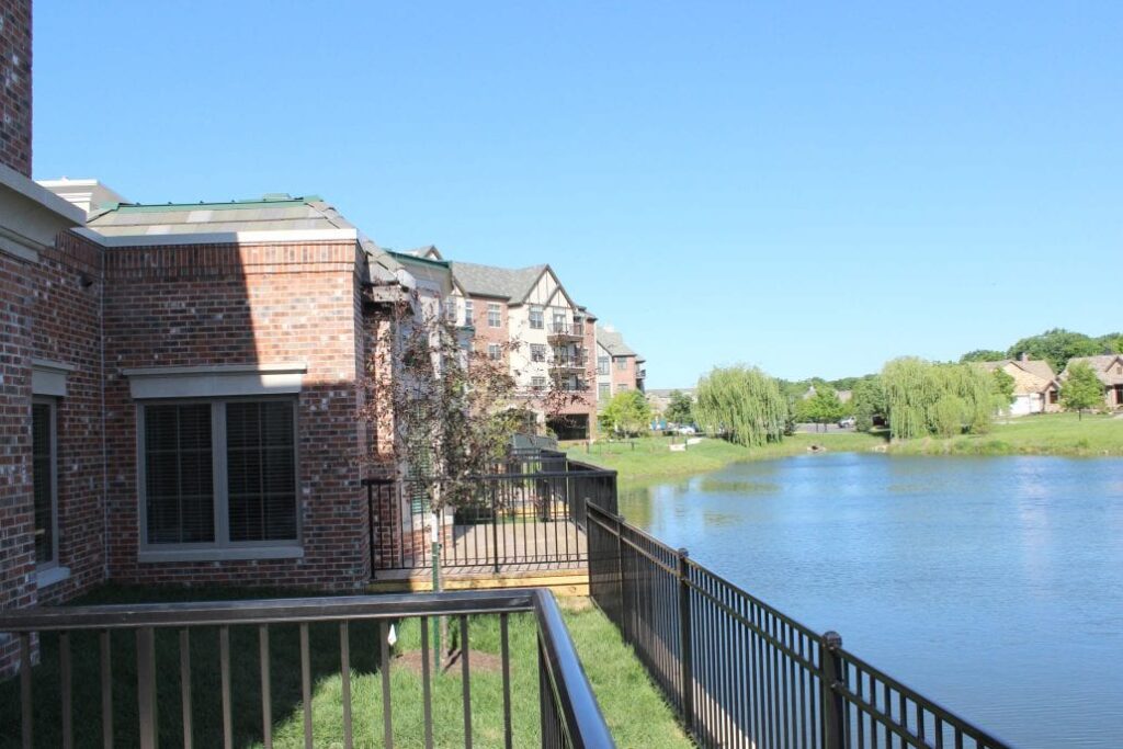 View from a balcony showing red-brick residential buildings beside a calm lake with trees and landscaped surroundings.