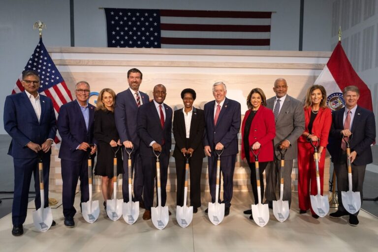 Group of men and women in business attire stand in a row holding ceremonial shovels, with American and Missouri flags behind them.
