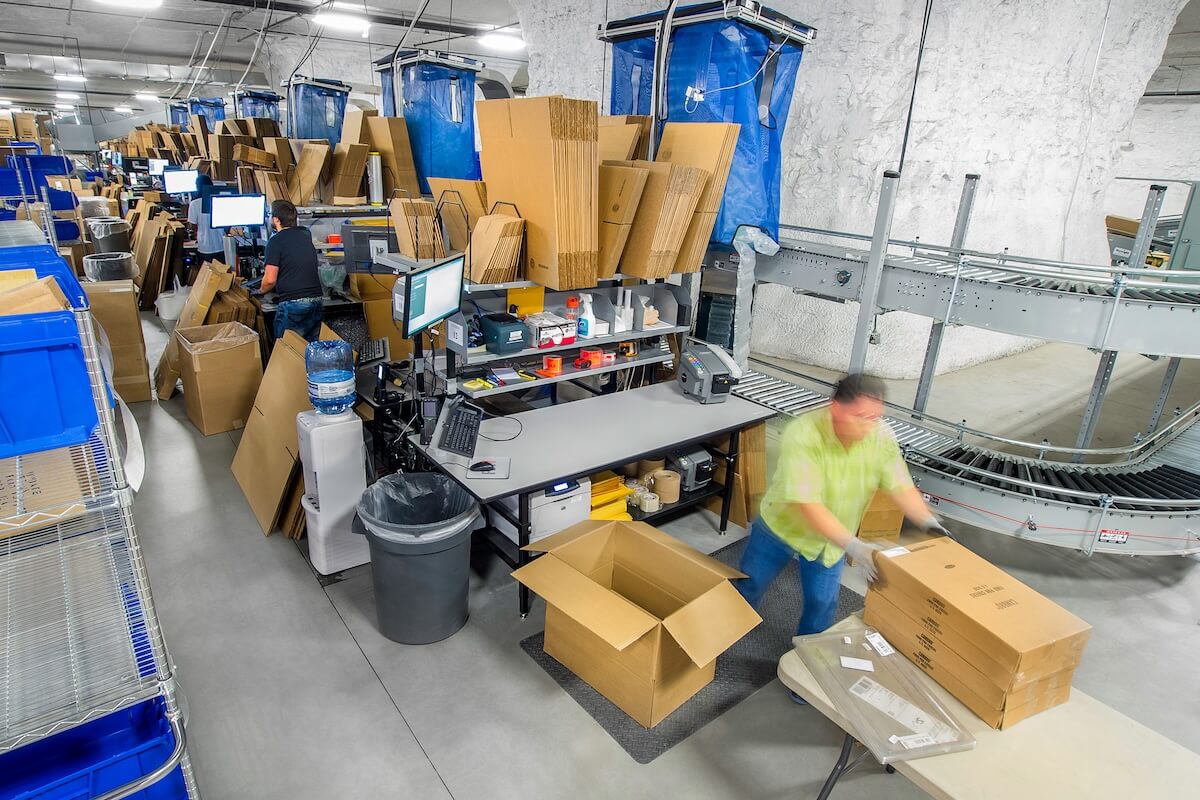 Workers at a packaging station surrounded by cardboard boxes, monitors, and supplies prepare shipments. A conveyor belt runs nearby, with one worker folding boxes and another handling flat-packed cartons.