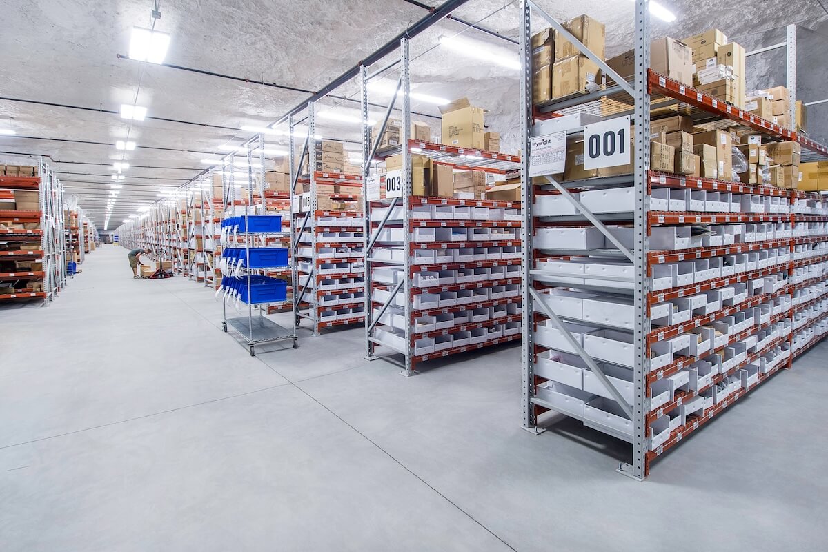 Rows of tall metal shelving units filled with white bins and cardboard boxes extend deep into a brightly lit underground warehouse. Signs with numbers label each section, and a worker in the distance is handling items near a pallet.