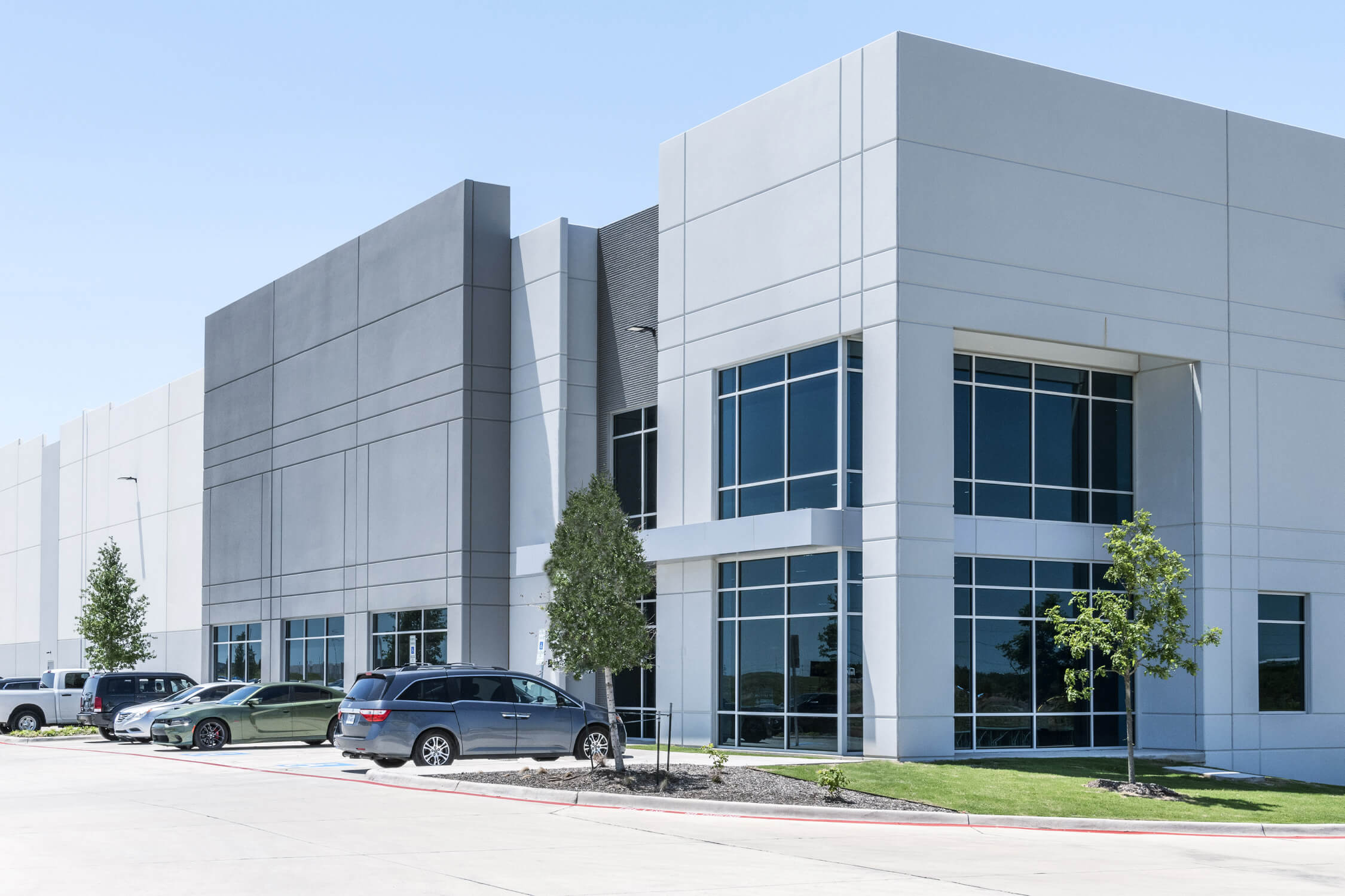 Contemporary industrial building featuring tall glass windows, smooth gray and white panel walls, and minimal landscaping with young trees. Several cars are parked along the concrete drive under a bright blue sky.
