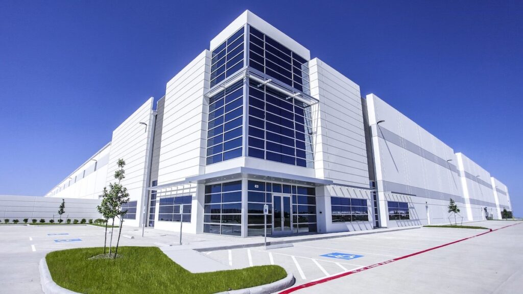 Large white industrial warehouse with a glass corner entry tower, clean lines, and an empty parking lot under a bright blue sky.