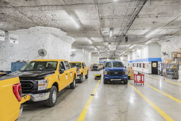 Bright underground garage-like space with yellow and blue pickup trucks parked in marked lanes beside storage and work areas.