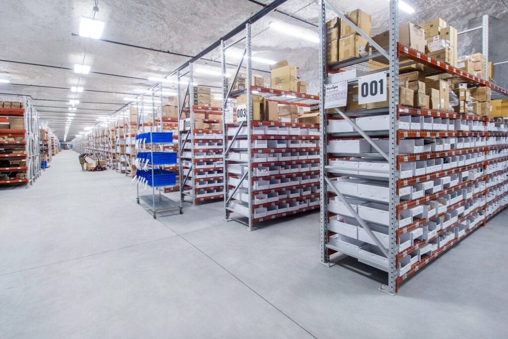 Brightly lit underground storage facility with tall metal shelves holding labeled boxes, cartons, and bins in neatly organized rows.