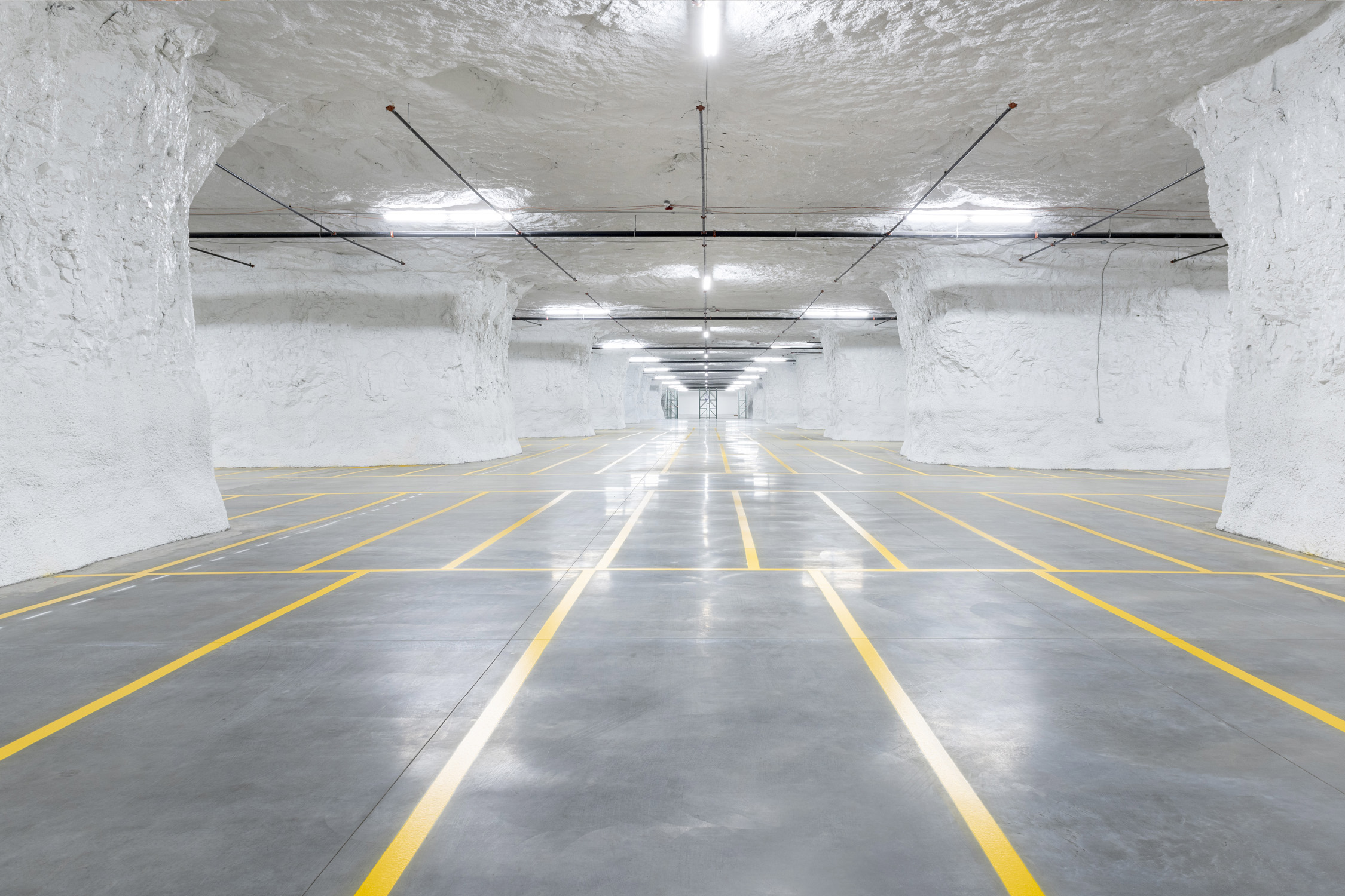 Clean, expansive subterranean facility with white rock walls and ceiling, smooth concrete floors marked by yellow lines, and evenly spaced fluorescent lights overhead.