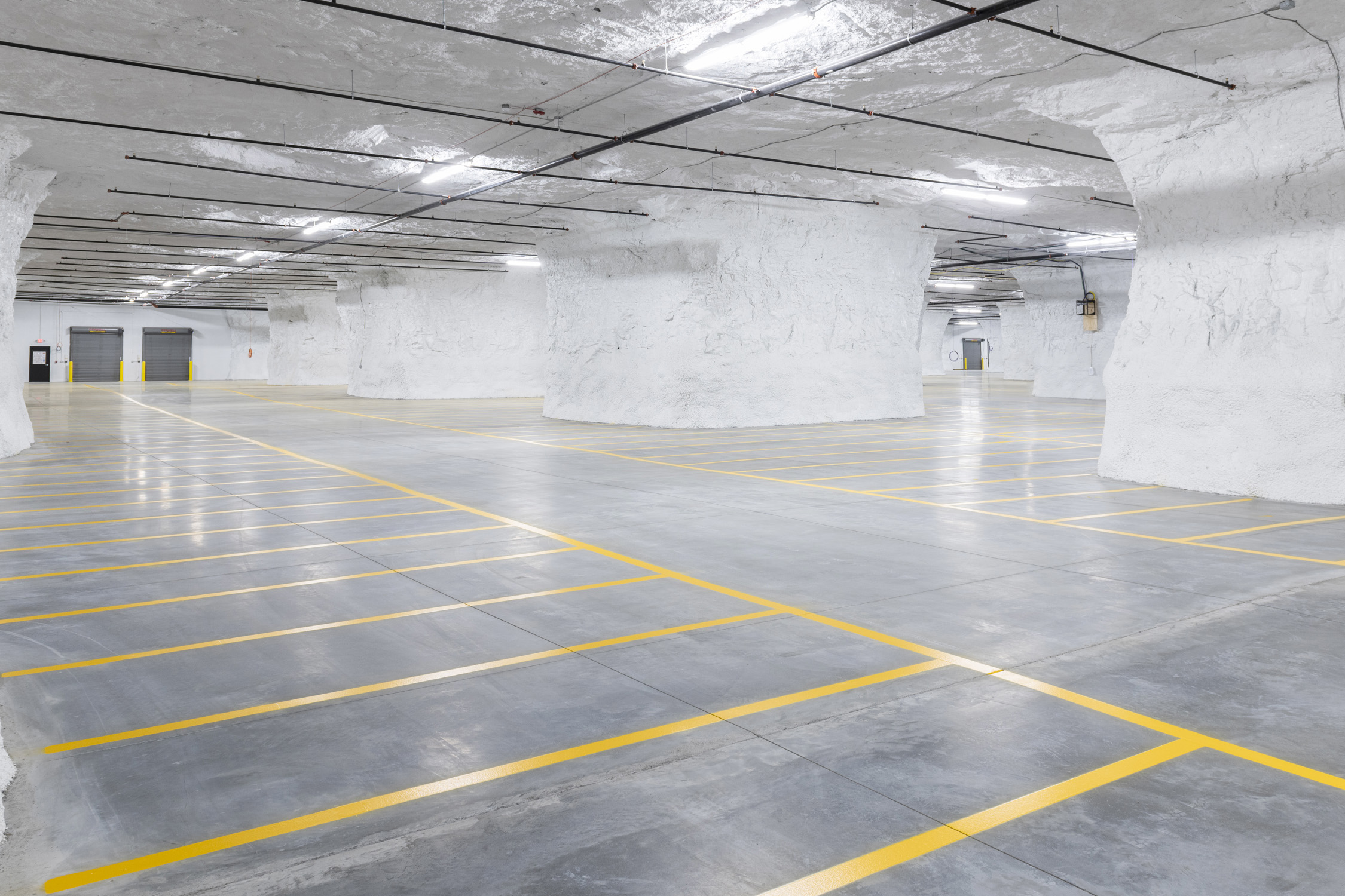 Spacious subterranean area featuring smooth gray concrete floors marked with yellow lines, large white limestone columns, and overhead fluorescent lighting.